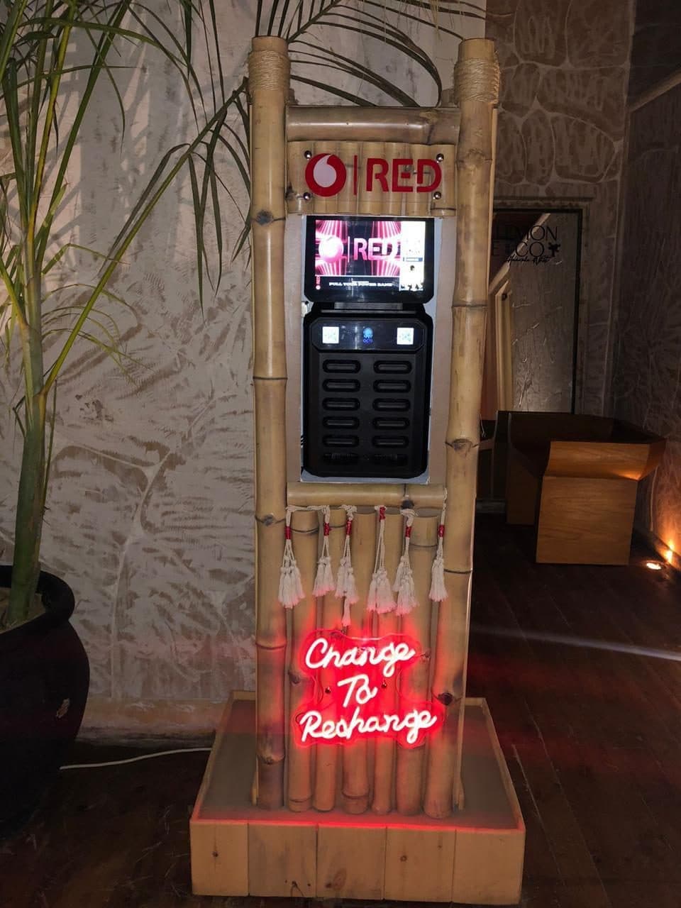 A bamboo charging station indoors at night, lit by a pink neon sign reading Change To Recharge