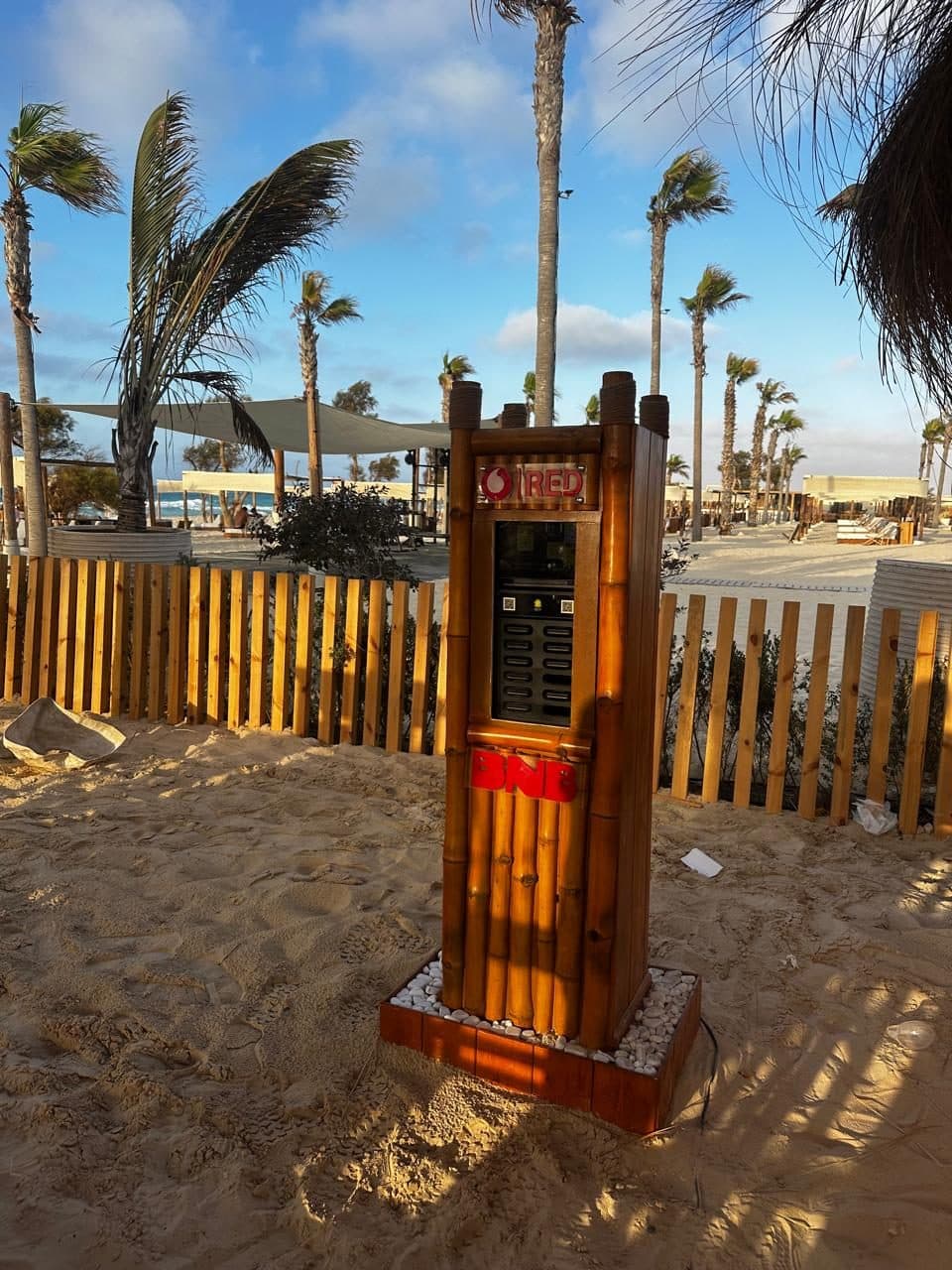 A charging station clad in bamboo standing on sand at a beach club, palm trees bending in the wind behind it
