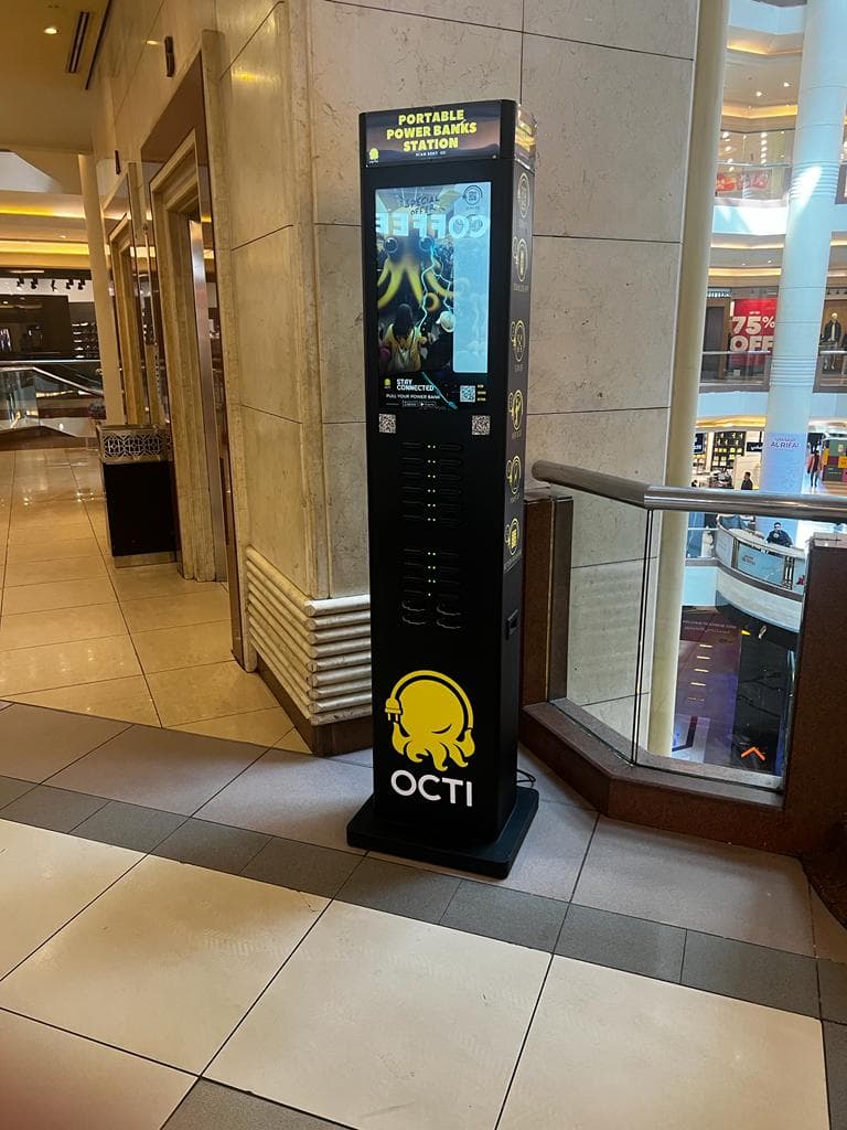 A charging station standing in a marble mall atrium beside a balustrade, shop fronts on the level below